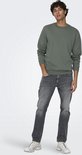 Man in groene crewneck trui en grijze jeans, handen in zakken.