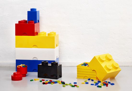Stapel van LEGO opbergdozen in rood, geel, wit en blauw, met een zwarte opbergdoos en gekleurde LEGO blokjes op de vloer.