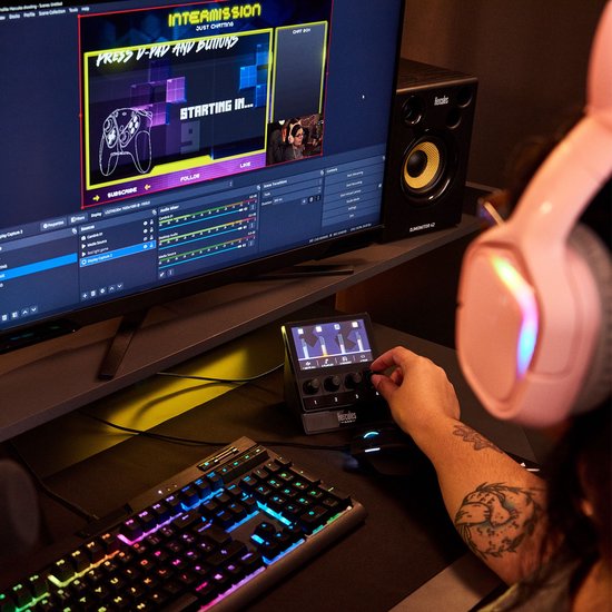 Een persoon met een roze koptelefoon gebruikt een zwarte audiocontroller op een bureau met een gamingtoetsenbord en dubbele monitoren.