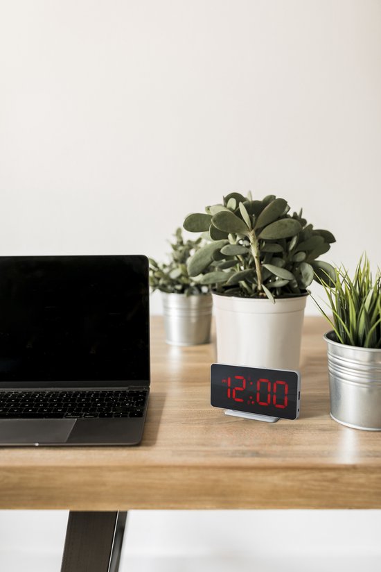 Witte digitale wekker met rood display dat 12:00 toont, op een houten bureau naast een laptop en planten.