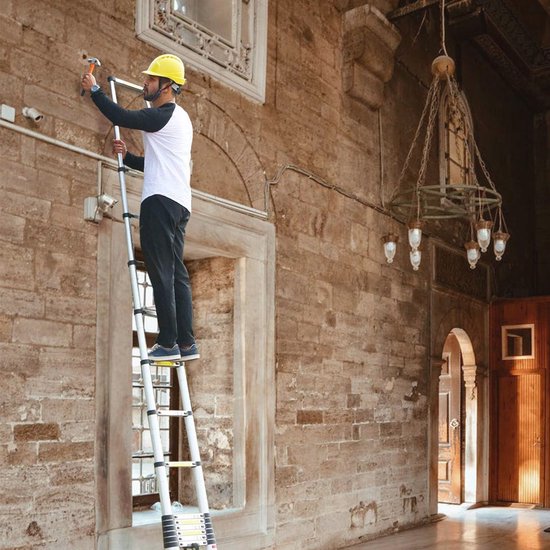Man met helm staat op een aluminium telescopische ladder en slaat een spijker in een stenen muur.