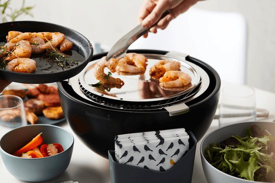 Zwarte tafelgrill met gegrilde garnalen, een hand legt er met een tang nog meer op.