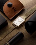 Gouden rechthoekig horloge met mesh band, omringd door bruine leren etui en zwarte accessoires op hout.
