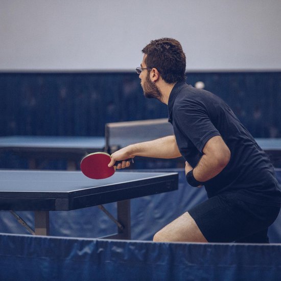 Man met bril en zwarte polo slaat tafeltennisbal met rode tafeltennisbat aan blauwe tafel.