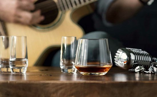 Deux verres à whisky en cristal remplis, sur une table en bois, avec un guitariste et un microphone en arrière-plan.