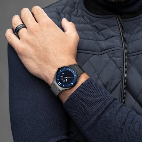 Man met zwart horloge om de pols. Blauwe wijzerplaat met zilveren indexen en datum. Zwarte Milanese band.