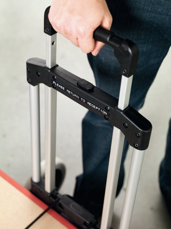 Hand met spijkerbroek pakt de metalen handgreep van een trolley met de tekst "PLEASE RETURN TO RECEPTION".