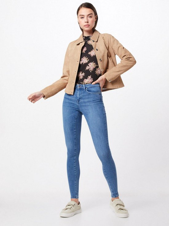 Model draagt een blauwe skinny jeans met middelhoge taille, met een beige jasje en bloemen top.