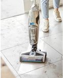 BISSELL Crosswave C3 Select stofzuiger met dweil op een vuile tegelvloer, met een persoon op de achtergrond.