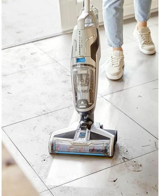 BISSELL Crosswave C3 Select stofzuiger met dweil op een vuile tegelvloer, met een persoon op de achtergrond.