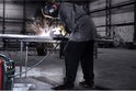 Rood lasapparaat op de voorgrond, terwijl een persoon een metalen pijp last in een grote werkplaats.