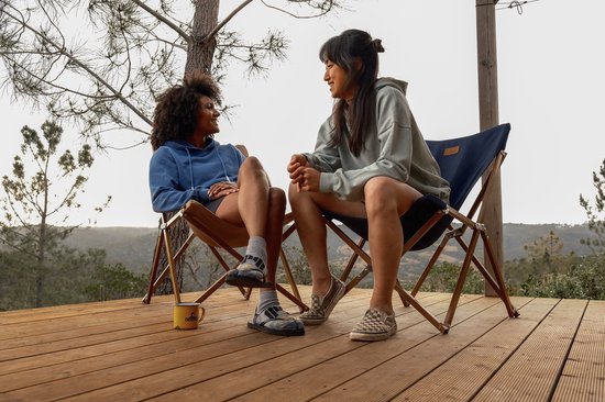 Twee vrouwen in hoodies zitten op kampeerstoelen op een houten terras in de natuur.