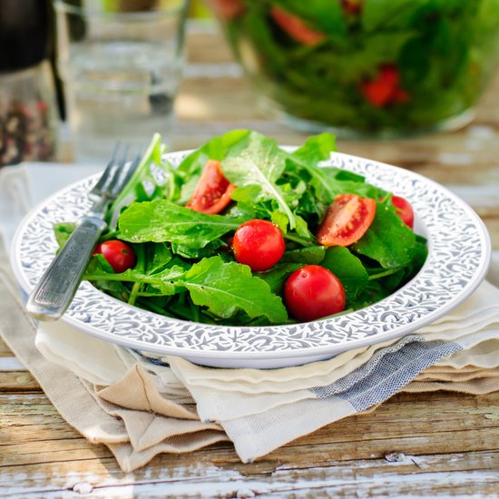 Een kom met rucola en cherrytomaatjes, versierd met een zilverkleurig patroon op de rand. Een vork ligt ernaast.