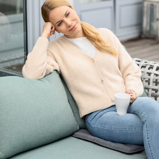 Een vrouw met spijkerbroek en vest zit ontspannen op een bank en houdt een mok vast. Een grijs kussen ligt op de bank.