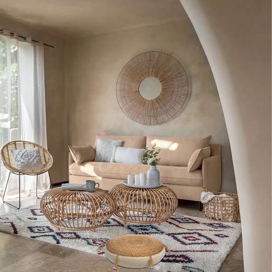 Un salon de couleur sable avec des tables en rotin, un canapé beige, un miroir rond et un pouf rond en jonc avec des pompons.