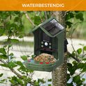 Groene vogelvoederhuis met zonnepaneel, hangend aan een boom in de regen, met "WATERBESTENDIG" tekst.