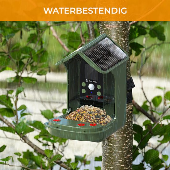 Groene vogelvoederhuis met zonnepaneel, hangend aan een boom in de regen, met "WATERBESTENDIG" tekst.