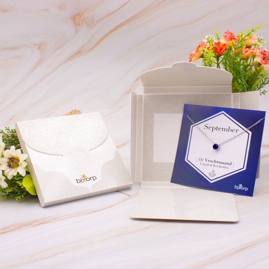 Zilverkleurige ketting met blauwe hanger, in een open doosje met bloemen.