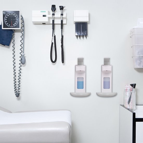 Een medische onderzoeksruimte met twee witte zeepdispensers aan de muur, een onderzoekstafel en medische apparatuur.