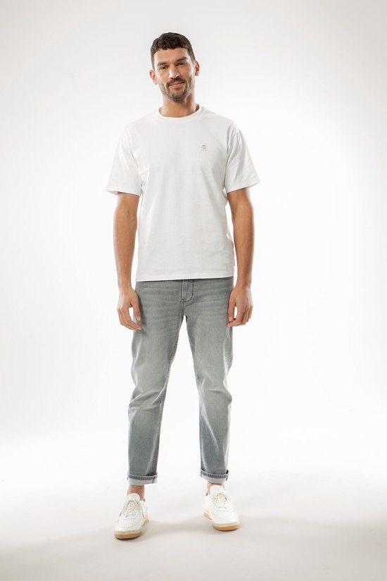 Man in een witte t-shirt en grijze jeans, staand met handen in de zakken.
