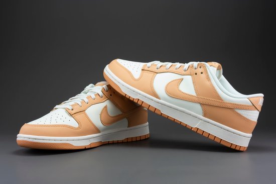 Een paar Nike Dunk Low damesschoenen, wit met oranje overlays, op een grijze achtergrond.