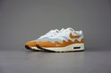 Wit met oranje Nike Air Max 1 Patta sneakers met beige en bruine accenten, op een grijze achtergrond.