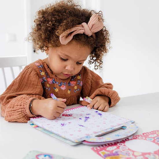 Une jeune fille aux cheveux bouclés et un bandeau, dessinant avec concentration dans un livre d'activités Licorne.
