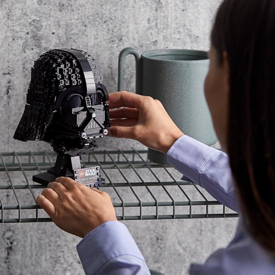 Een vrouw plaatst een LEGO-helm van Darth Vader op een displaystandaard, met een grijze mok op de achtergrond.