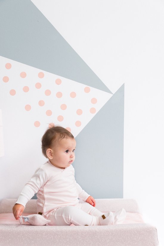 Un bébé est assis sur une surface rose, portant une tenue rose pâle. En arrière-plan, un mur présente des motifs géométriques et des pois roses.
