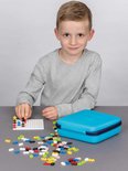 Een jongen zit aan tafel en bouwt met Plus-Plus speelgoedblokjes op een wit pegboard, naast een turquoise koffer.