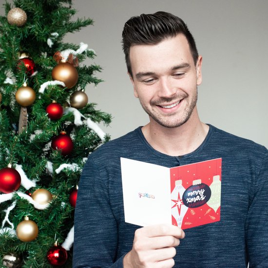 Man met kerstkaart met kerstballen, tekst 'merry xmas'. Achtergrond: kerstboom met ornamenten.