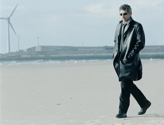Homme en long manteau noir et lunettes de soleil marchant sur la plage, avec des éoliennes en arrière-plan.