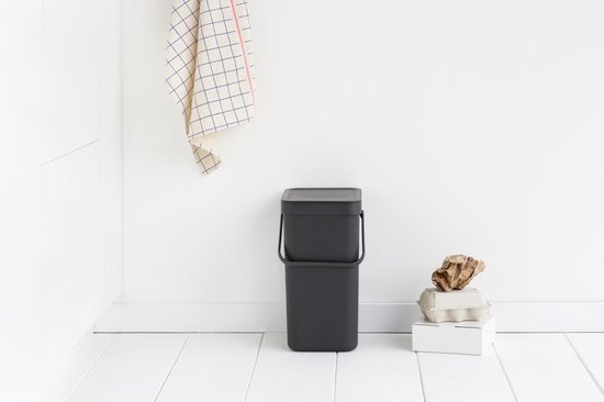 Poubelle Brabantia gris foncé avec couvercle entrouvert, dans une pièce blanche avec décoration.