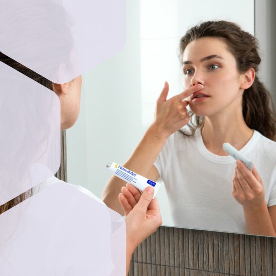 Vrouw kijkt in spiegel en doet NasuMel zalf op haar neus. Tube in linkerhand.