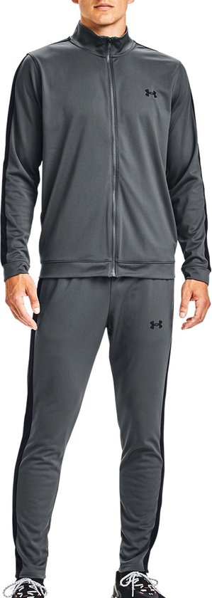 Man met een grijs Under Armour trainingspak. Zwarte strepen op de mouwen en zijkanten van de broek. Zwarte logo's.