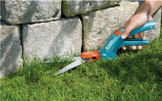 Hand gebruikt blauwe Gardena graschaar om gras te knippen bij stenen muur.