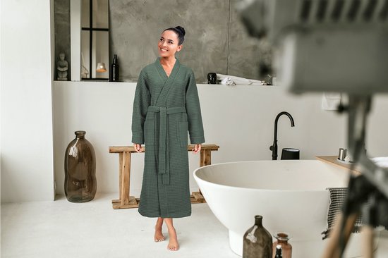 Vrolijke vrouw in een olijfgroene wafel badjas in een badkamer met een ligbad.