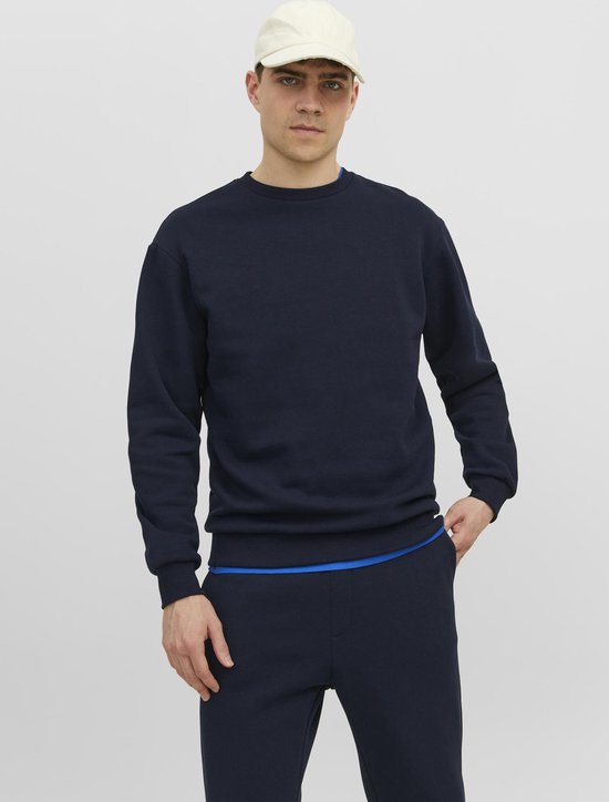 Man met witte pet, draagt een donkerblauwe effen sweater met ronde hals en blauwe voering. Daaronder een donkerblauwe broek.
