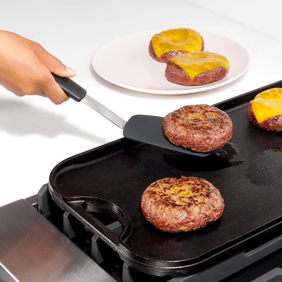 Hand met spatel die een cheeseburger van een grill verplaatst. Twee andere burgers met kaas op een bord.
