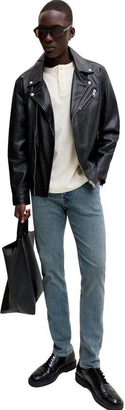 Man met zwarte bikerjas, crèmekleurige top, blauwe slim fit jeans, zonnebril en zwarte schoenen.