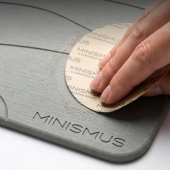 Main ponce un tapis de bain en diatomite gris avec logo "MINISMUS". Petites particules de poussière visibles.