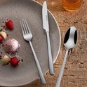 Gepolijste roestvrijstalen mes, vork en lepel op een bord met dessert, naast een glas.