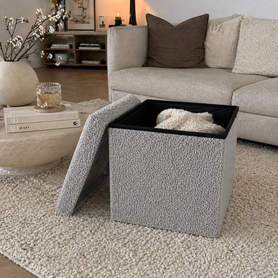 Grijze poef met teddy bouclé stof en zwart interieur, open met deken erin. In een woonkamer.
