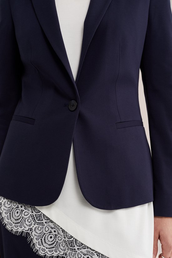 Sluiting van een donkerblauwe dames blazer, met een knoop en een witte top met kanten rand.