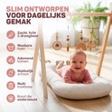 Vrolijke baby in roze outfit in een crèmekleurig babynestje onder een houten speelboog. Iconen voor zacht, wasbaar, ademend, makkelijk schoon, multi-inzetbaar, rand.
