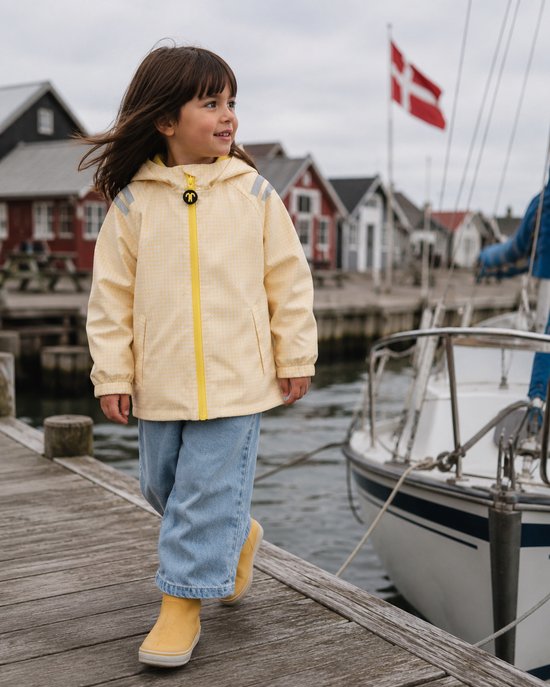 Meisje in lichtgele geruite regenjas op een houten pier bij een boot en huizen.