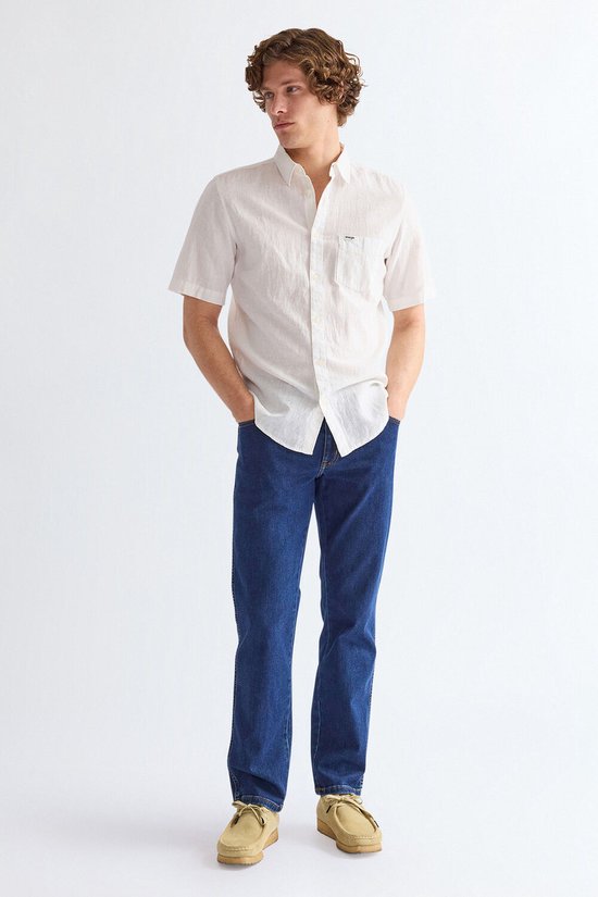Man met handen in zakken, draagt een wit linnen overhemd met korte mouwen en een donkerblauwe jeans.