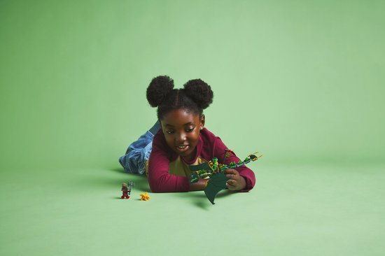 Jong meisje met afro-kapsel ligt op de grond en speelt met een groene LEGO draak en figuurtjes.