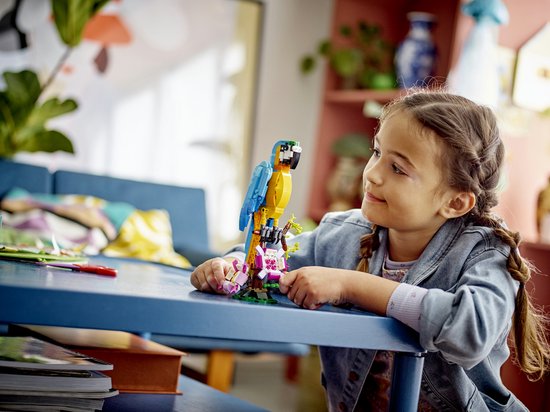 Een meisje met vlechtjes kijkt naar een gele en blauwe papegaai van LEGO-blokjes op een tak met roze bloemen.