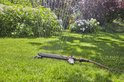 Grijze zwenksproeier spuit water op een groen gazon met bloeiende planten.